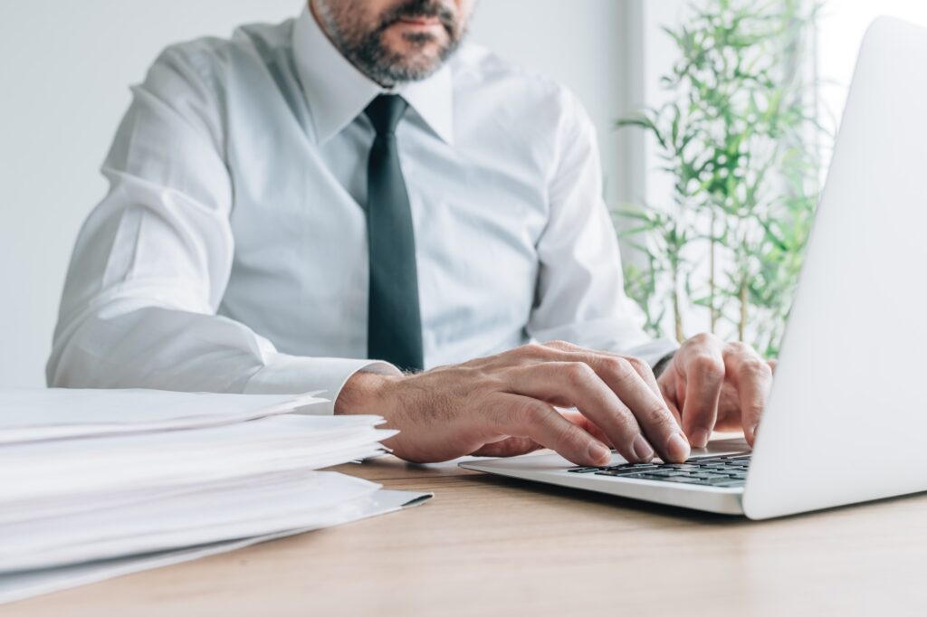 Businessman working on laptop computer with paperwork pile on office desk